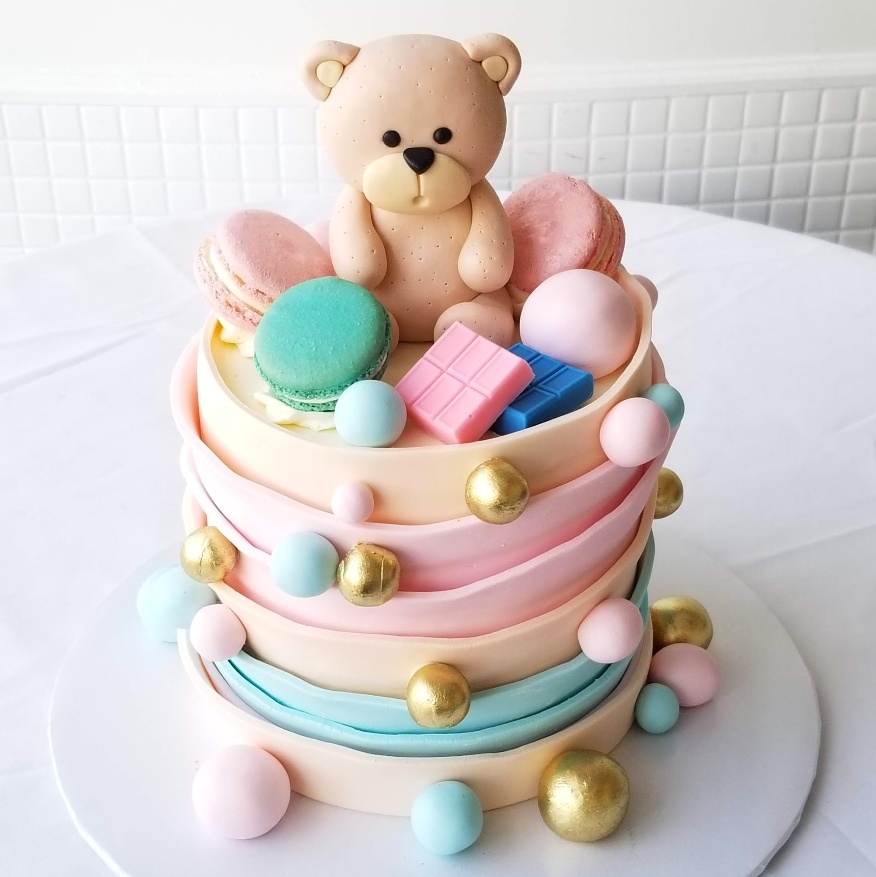 A pink and blue cake with a bear and macaron toppers